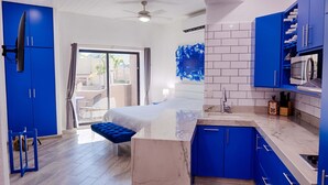 A modern hotel room with a large bed, a balcony with a view, a kitchen area, and blue cabinetry.