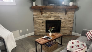 A living room with a stone fireplace, a television mounted above it, a sofa with floral cushions, and a coffee table with magazines. - PROMO 5% WEEKLY DISCOUNT, ONLY FOR  NOV/ DEC /JAN /FEB.
Downtown Jasper Suite (Jasper)