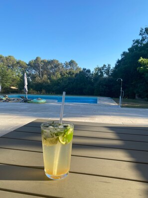 A glass of iced drink with mint and lime on a wooden table by a pool with a clear blue sky.