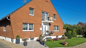 A two-story brick house with a balcony, a satellite dish, and a well-maintained garden with potted plants and flowers. - Vacation apartment Am Damm with a direct view of the Rhine DTV 4 stars (Wesel)