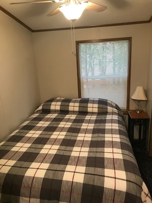 Quarto com cama, ventilador de teto, janela com persianas e mesa de cabeceira com abajur.