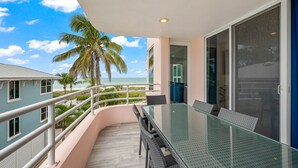 A balcony with a glass table and chairs, overlooking a beach with palm trees and a clear sky. - Beachfront Condo! Gulf Views, Pool, and Balcony (Holmes Beach)