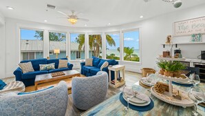 A spacious living room with a blue sofa and armchairs, a dining table set for a meal, and large windows offering a view of the ocean. - Beachfront Condo! Gulf Views, Pool, and Balcony (Holmes Beach)