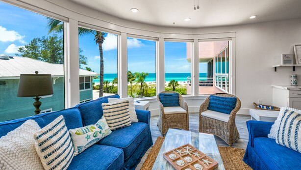 A spacious living area with a large window offering a view of the beach and ocean. There are two blue sofas and wicker chairs with cushions. - Beachfront Condo! Gulf Views, Pool, and Balcony (Holmes Beach)