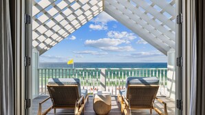 Een open balkon met twee rieten stoelen en een houten tafel, met uitzicht op een strand met een witte hek en een vlag.