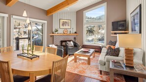 A cozy living room with a fireplace, a dining area, and large windows offering a view of the outdoors.
