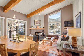 A cozy living room with a fireplace, a dining area, and large windows offering a view of the outdoors. - Woodbridge Condos by iTrip Aspen Snowmass (Snowmass Village)