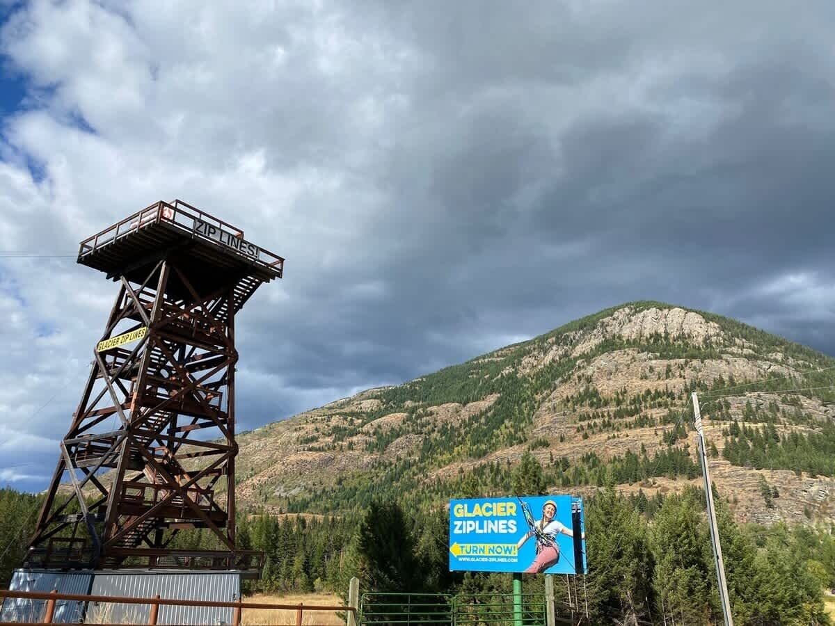 A metal tower structure with a sign for Glacier ZipLines, a mountain in the background, and a clear sky.