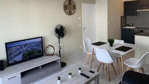 Ett modernt vardagsrum med en platt-TV, en bordsfläkt, ett matbord med stolar, en liten krukväxt och en köksdel med skåp och en spis.