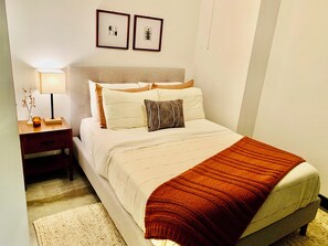 A neatly made bed with a white comforter and a brown knitted throw, a bedside table with a lamp and a candle, and two framed artworks on the wall.