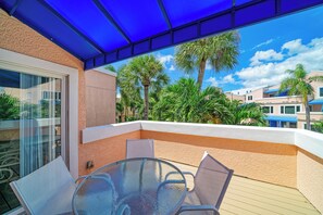 A covered outdoor seating area with a glass table and chairs, overlooking a landscaped garden with palm trees and a clear blue sky.