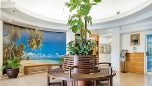 Eine Lobby mit einem runden Holztisch, zwei Stühlen, einer großen Topfpflanze, einer Bank und einem Wandbild mit einer Strandansicht.
