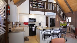 Cocina equipada con microondas, estufa y una isla con sillas. Se ve una escalera que sube a un altillo con estanterías.