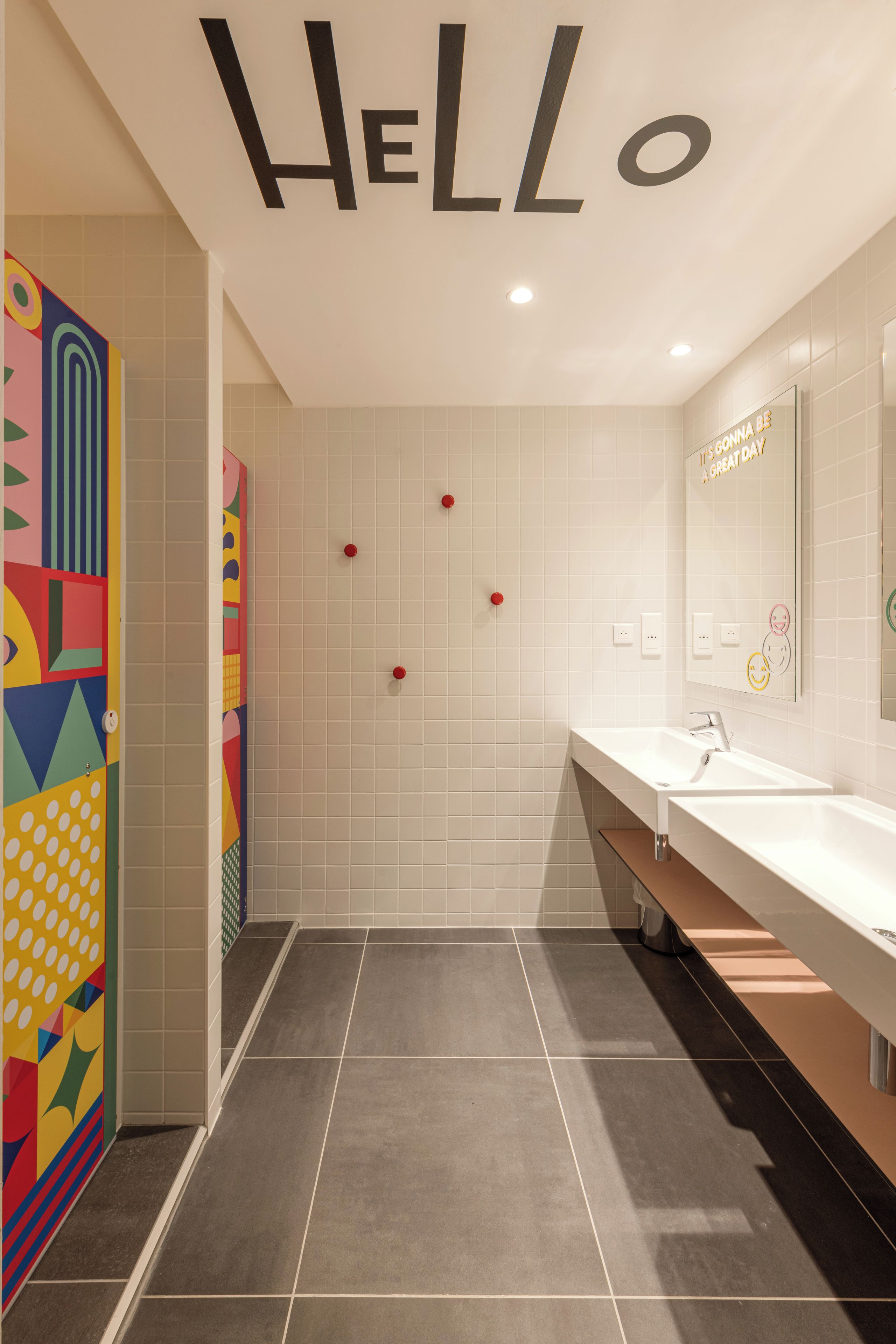 Une salle de bain moderne avec un panneau « BONJOUR » au plafond, un grand miroir et un motif géométrique coloré sur le mur.