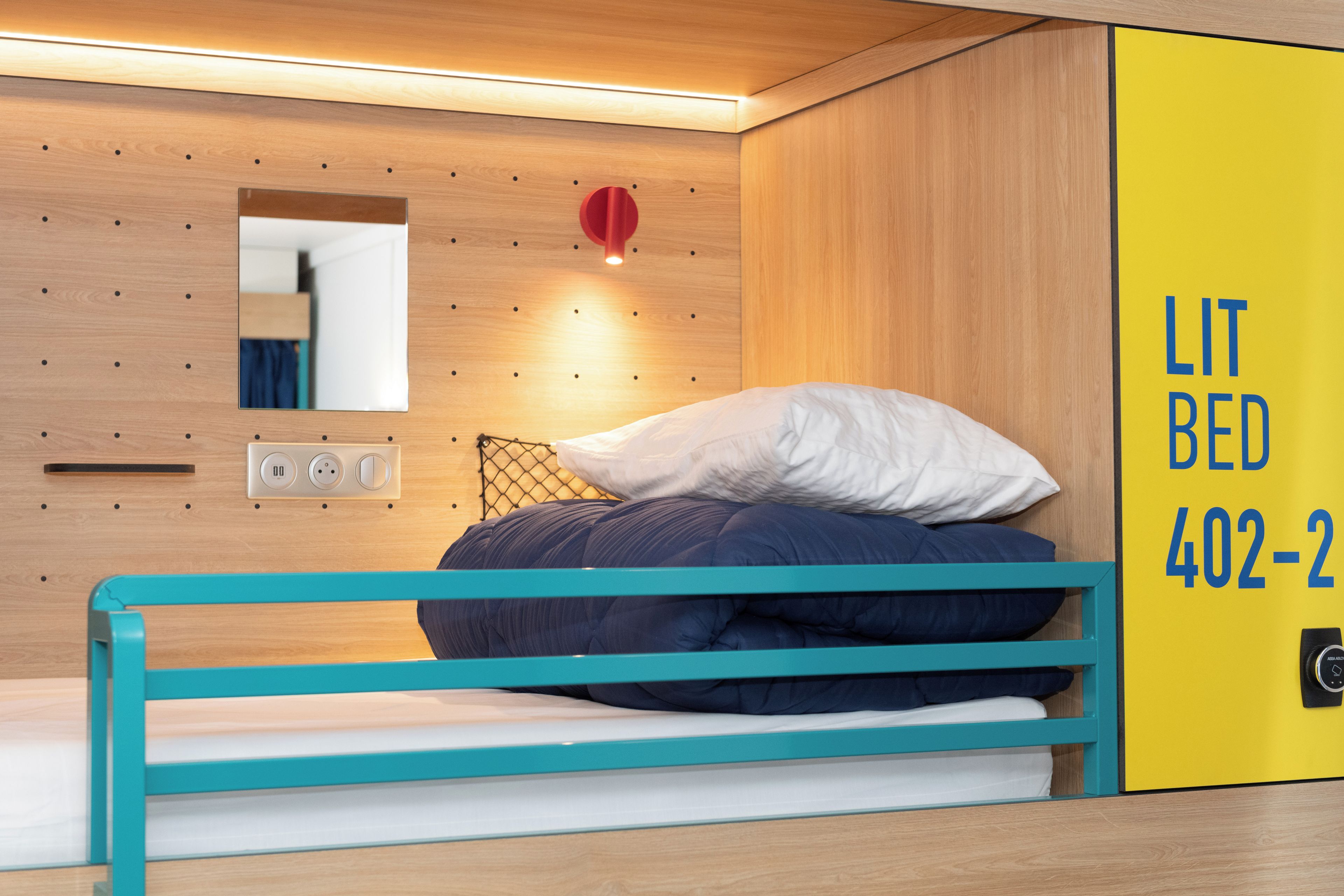 A compact room with a bunk bed, a small mirror, a wall-mounted light, and a yellow wall with a phone number.