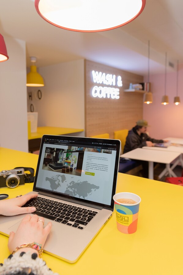 A person using a laptop at a yellow table with a cup of coffee, in a room with a 'WASH & COFFEE' sign.