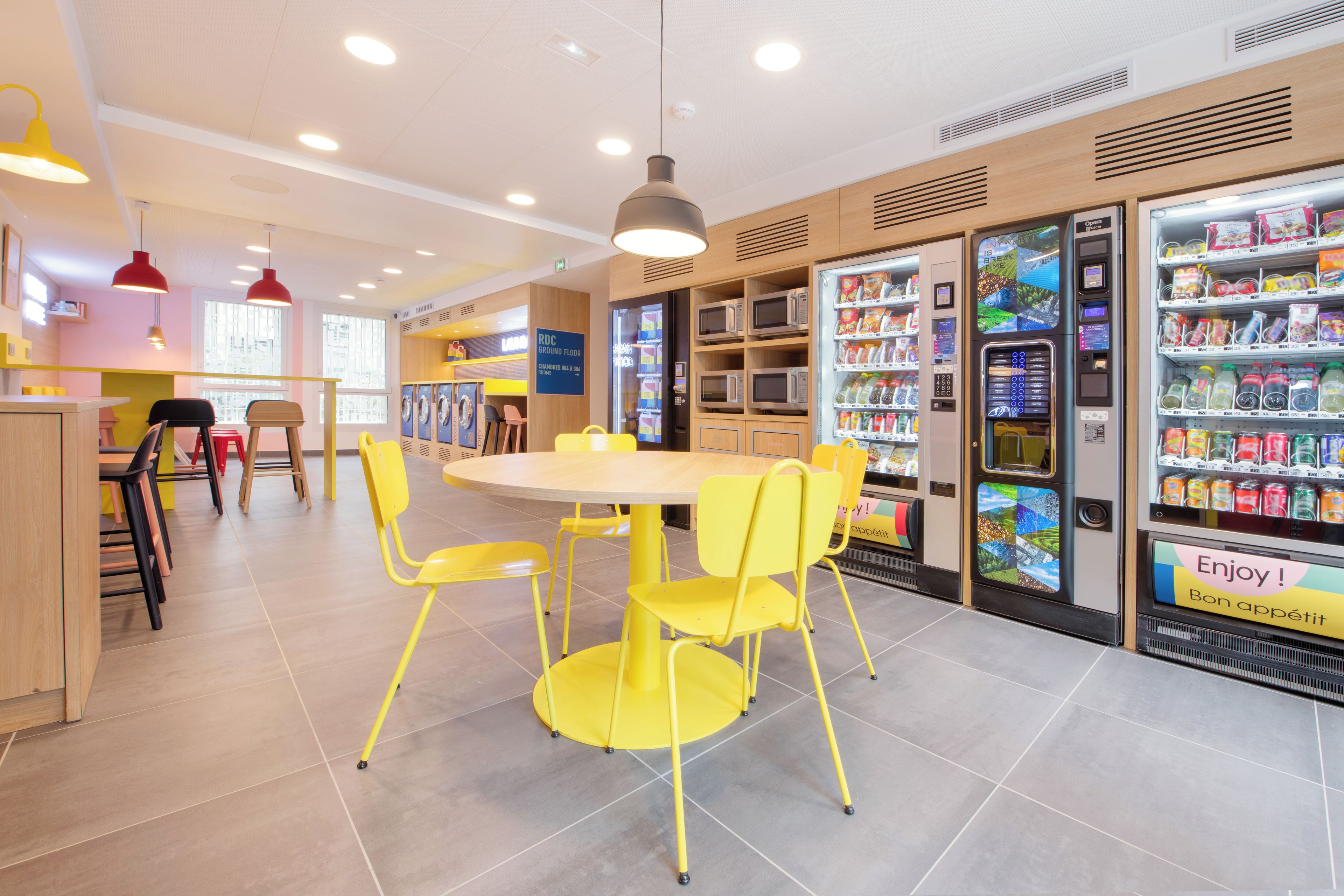 A brightly lit indoor space with a round table surrounded by yellow chairs, vending machines stocked with snacks and drinks, and a visible sign indicating a ground floor location.