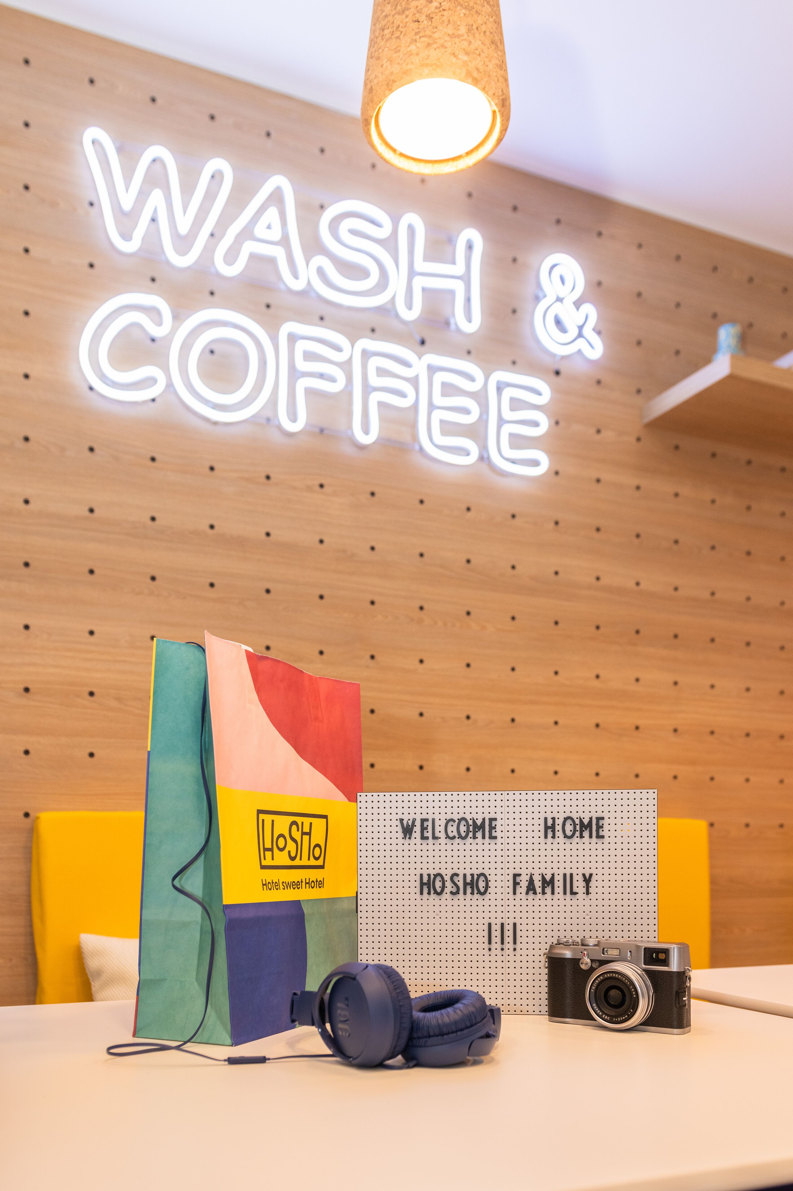 Una habitación de hotel con un letrero de neón que dice "WASH & COFFEE" y un cartel de bienvenida que dice "BIENVENIDOS FAMILIA HOSHO".