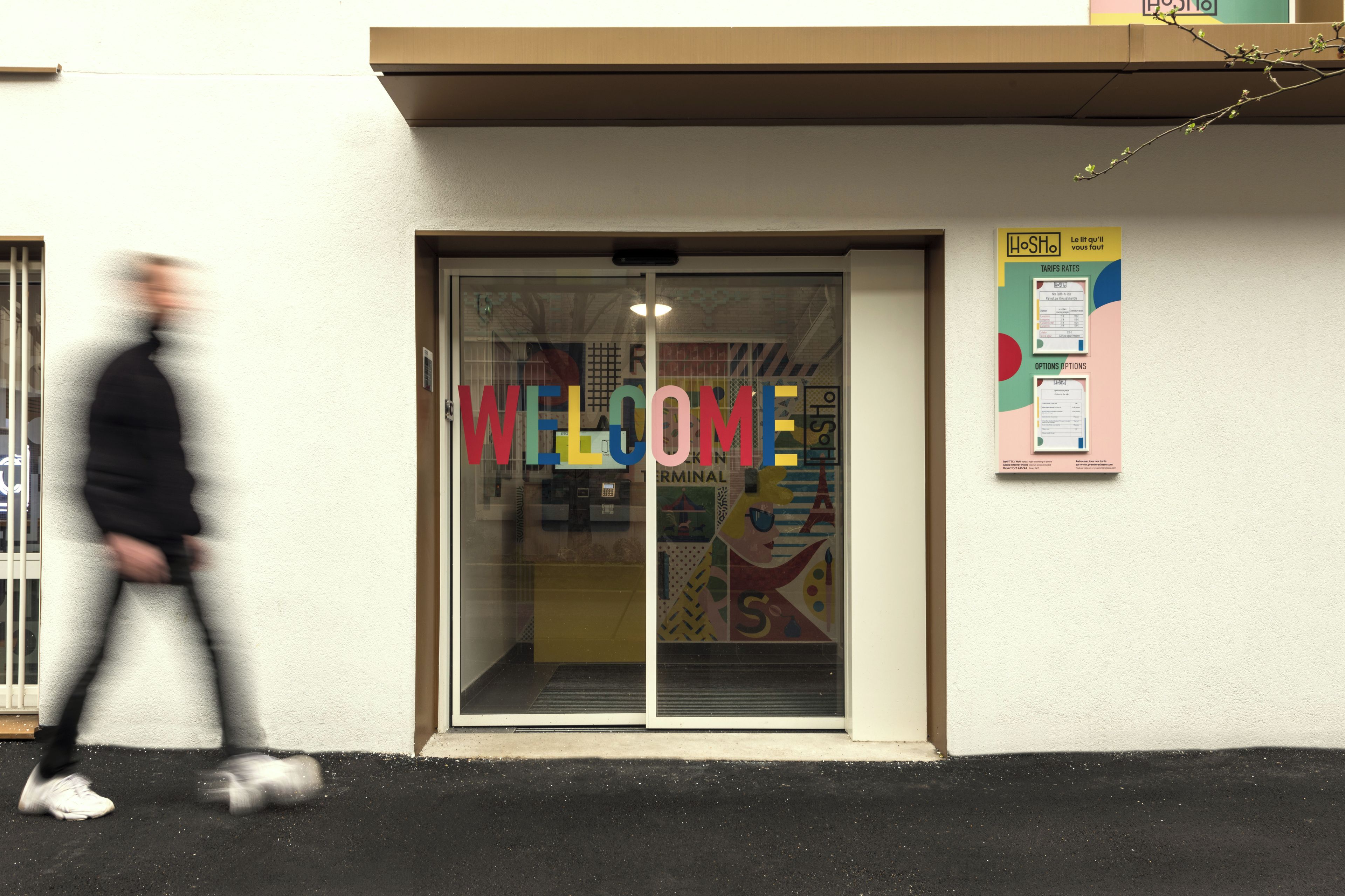 L’entrée d’un hôtel, avec une grande porte vitrée sur laquelle est affiché un panneau coloré avec le mot « BIENVENUE ».