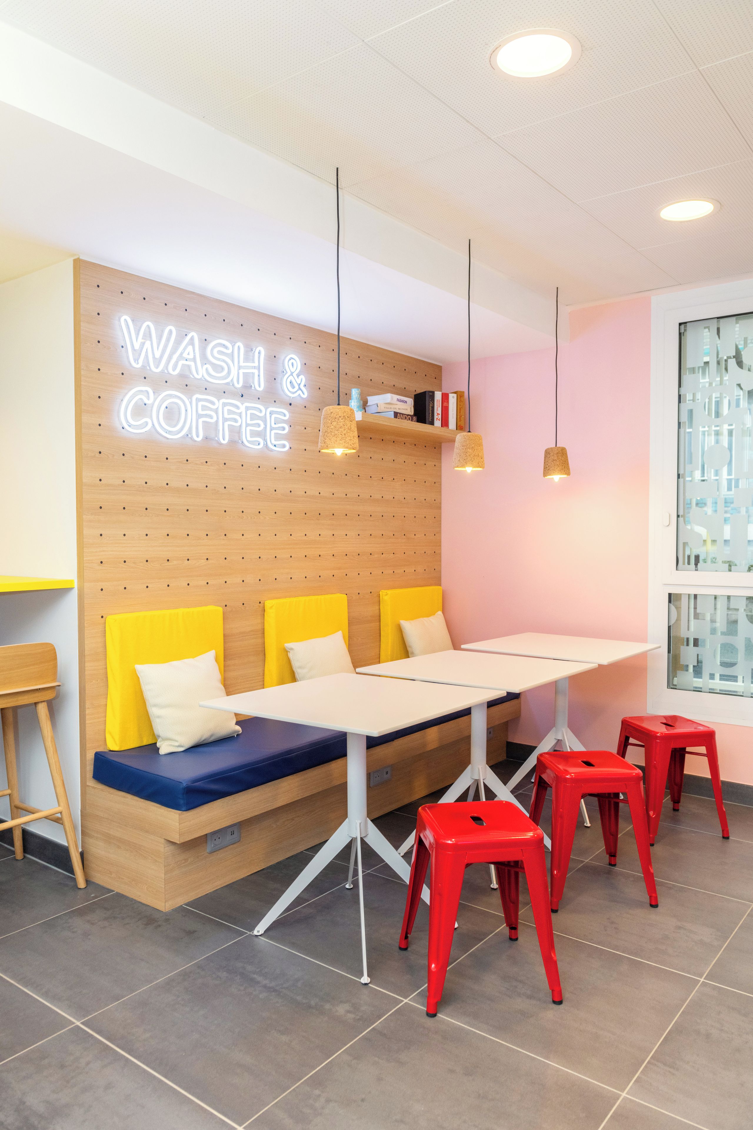 A modern cafe interior with a neon sign reading 'WASH & COFFEE', wooden wall panels, yellow cushioned seating, and red stools.