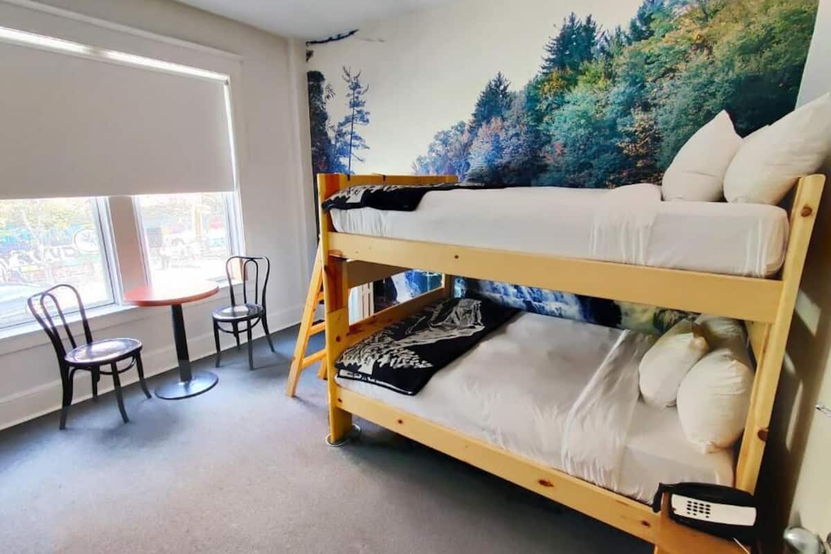 Un lit superposé avec une fresque représentant une forêt peinte sur le mur derrière, une petite table avec des chaises et une fenêtre avec des stores.