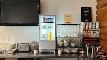 A kitchen area with a refrigerator stocked with beverages, a toaster, and a sign indicating breakfast availability.