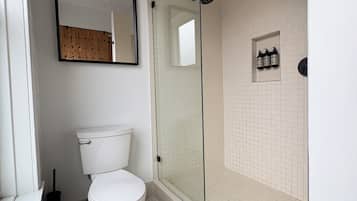 A bathroom with a toilet, a glass-enclosed shower, a mirror, and a small shelf with toiletries.