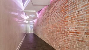 Un couloir avec un mur de briques d’un côté et un mur uni de l ’autre, qui mène à une pièce lumineuse dotée d’une grande fenêtre.