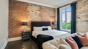 Une chambre avec un mur en briques, un grand lit, une table de chevet, une fenêtre avec des rideaux et une vue sur la verdure à l’extérieur.
