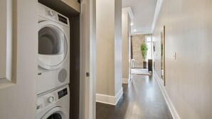 Une buanderie avec des lave-linge et des sèche-linge superposés, un couloir avec un mur en briques et une porte donnant sur une autre pièce.