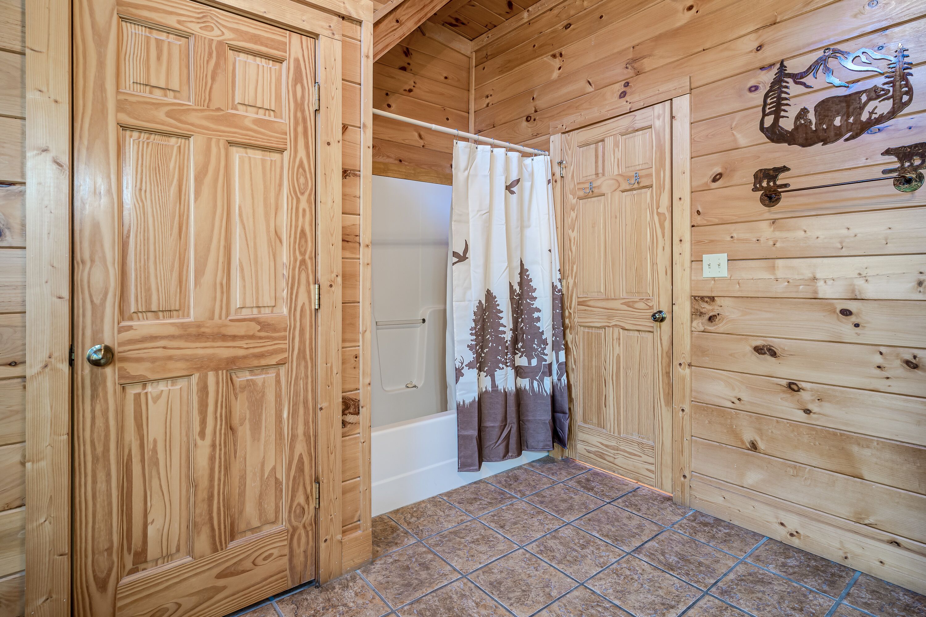 A wooden cabin interior with a bathroom featuring a shower curtain with a forest design, a bathtub, and a wooden door.