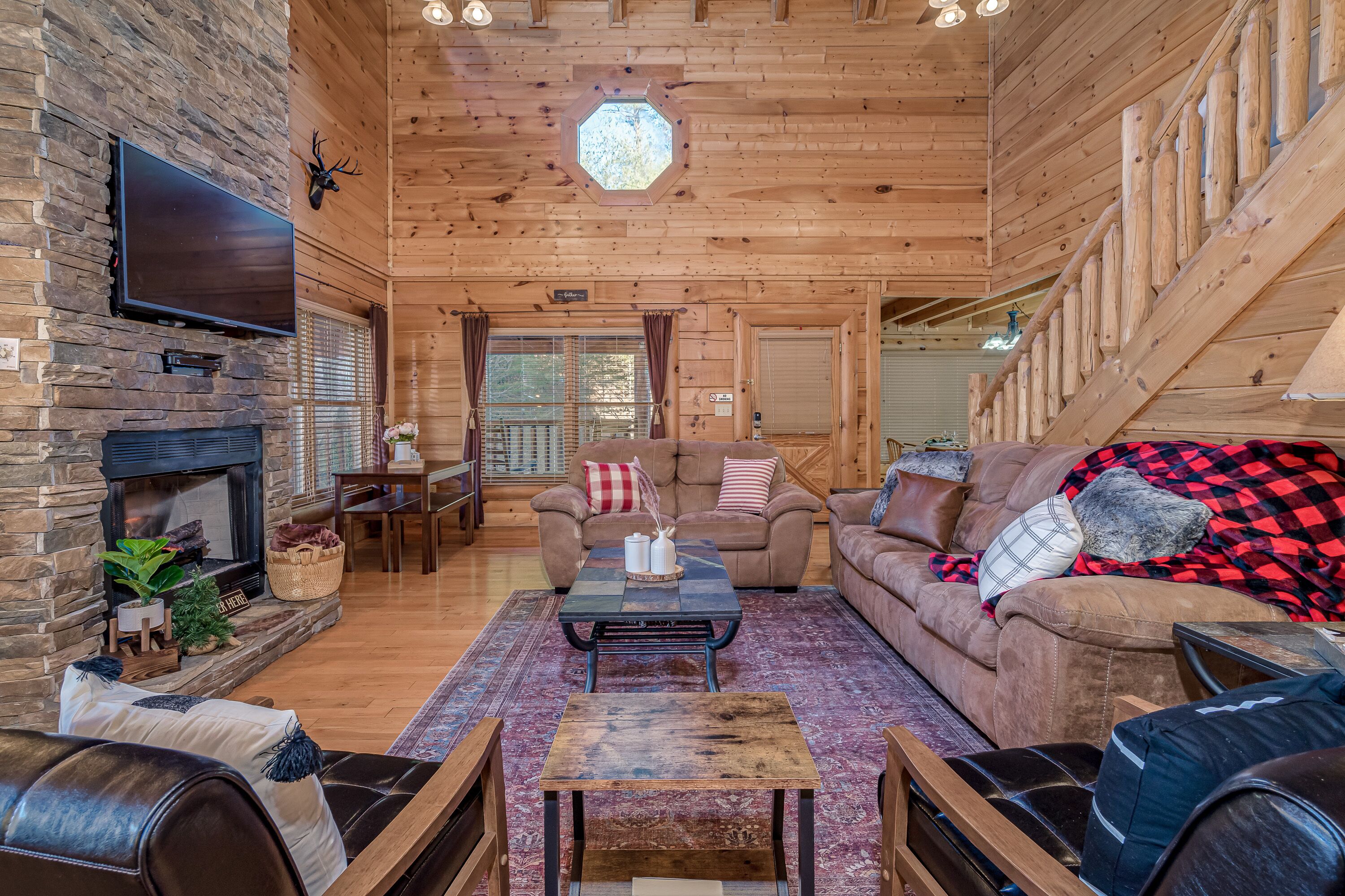 A cozy log cabin interior with a stone fireplace, a flat-screen TV, a wooden staircase, and a comfortable seating area with a couch and armchairs.