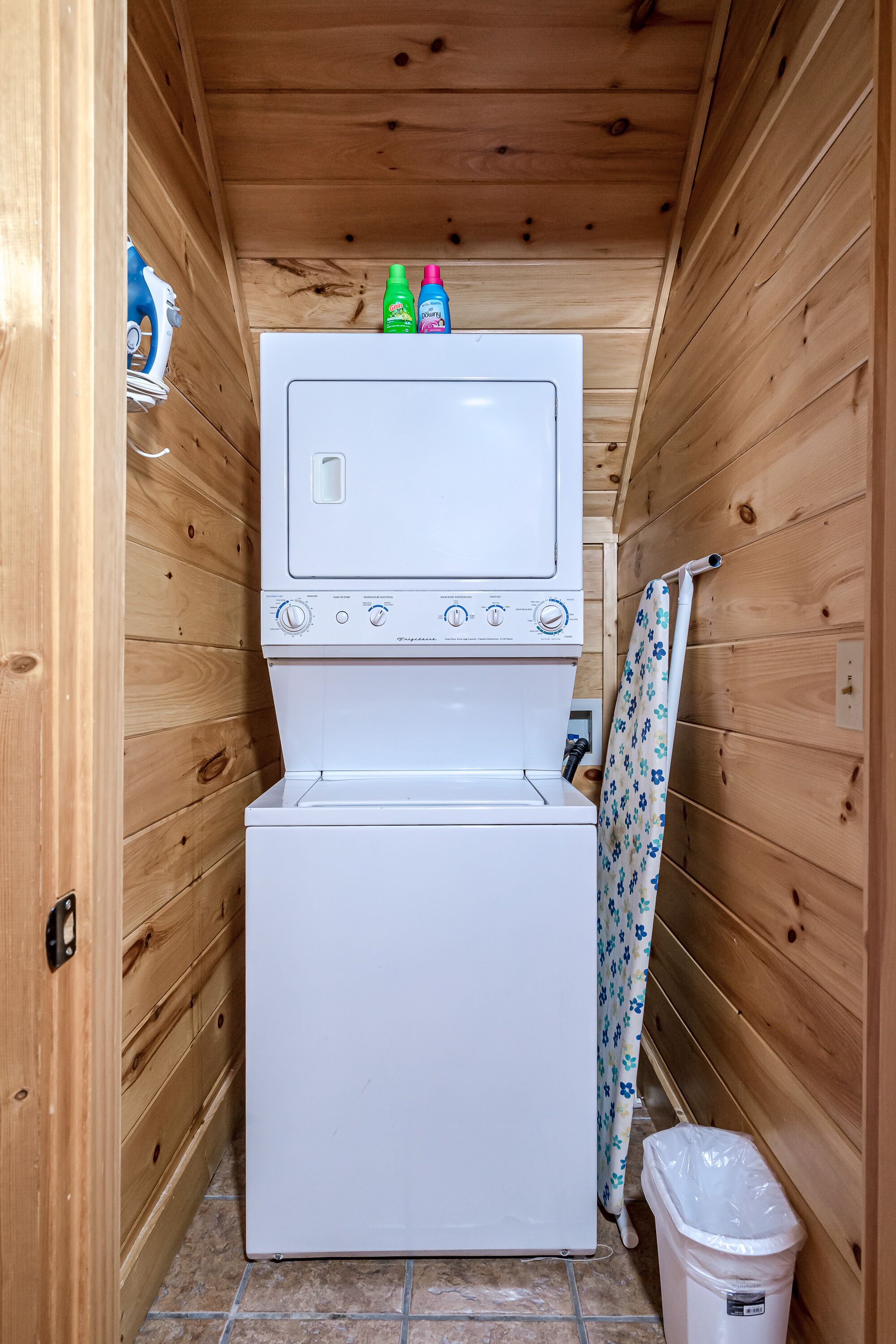 Een kleine wasruimte met een gestapelde wasmachine en droger, een klein afvalbakje en een douchegordijn.