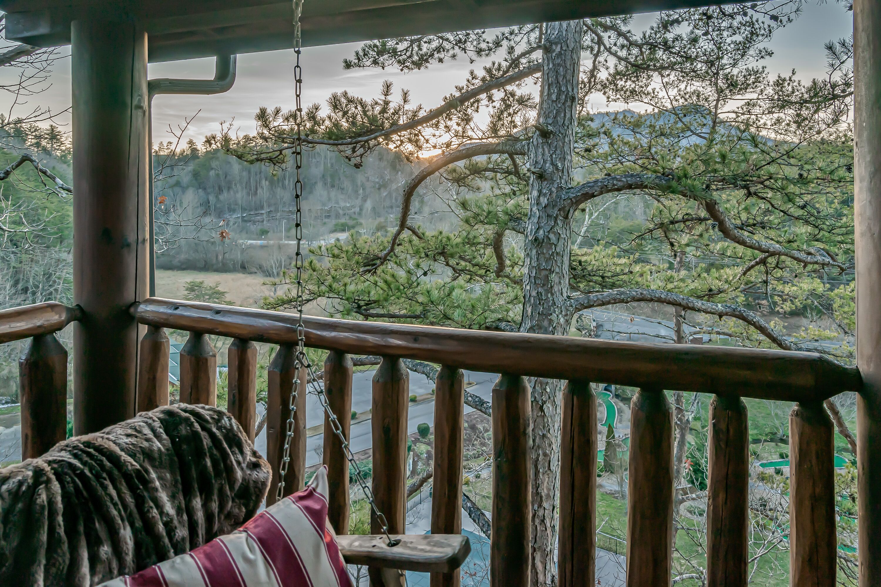 Een houten schommelstoel met een gestreept kussen op een veranda met uitzicht op een bergachtig landschap.