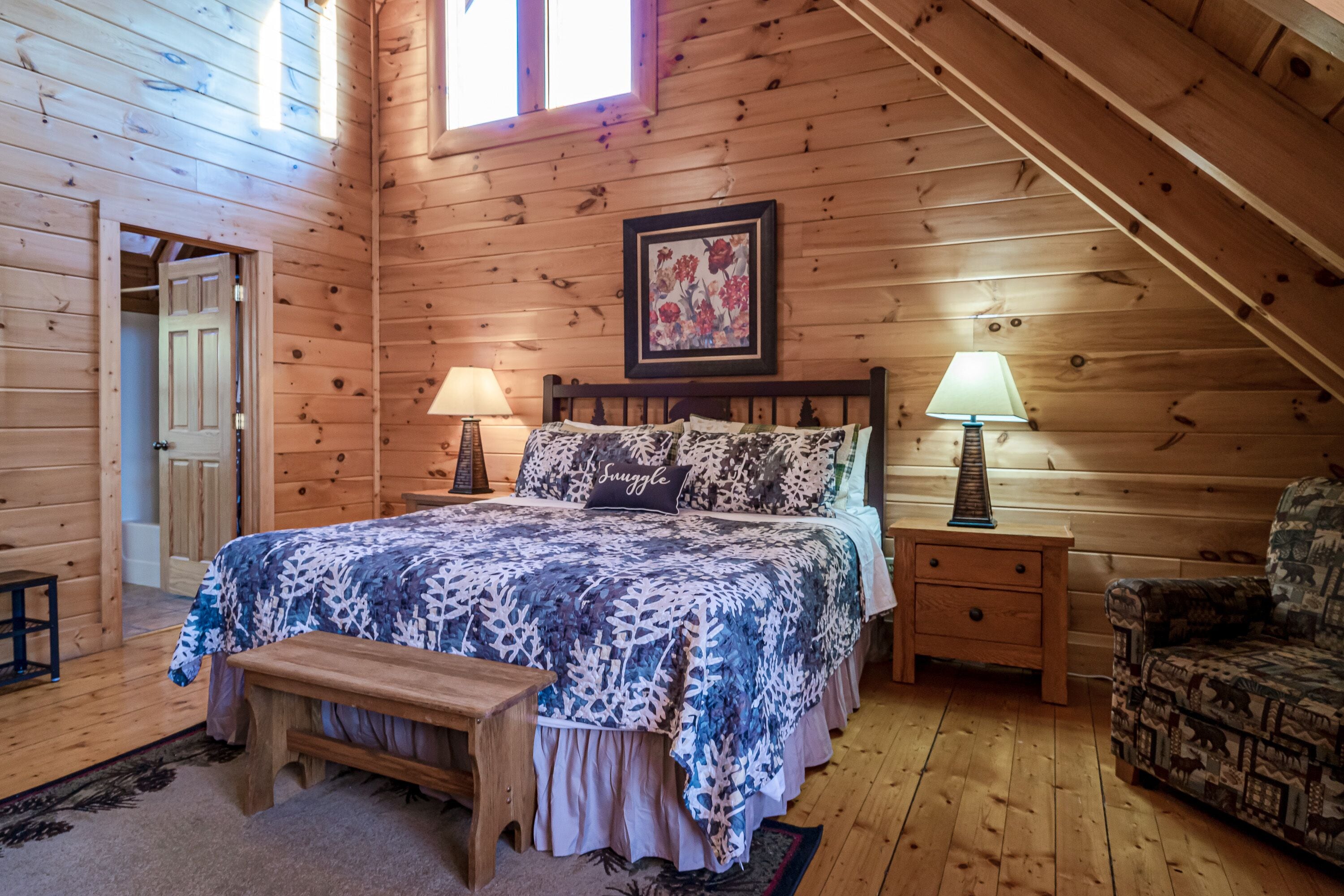 A wooden cabin bedroom with a bed, two bedside lamps, a bench, a chair, and a framed floral artwork on the wall.