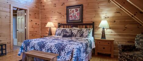 Une chambre dans une cabane en bois, avec un lit, deux lampes de chevet, un banc, une chaise et une œuvre d’art florale encadrée accrochée au mur.