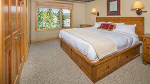 A bedroom with a large bed, wooden headboard, two bedside tables, a window with a view of trees, and a wall-mounted lamp. - Ski-In/Out Condo | Lone Peak Views & Community Pool/Hot Tub (Big Sky)