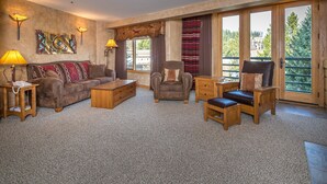 A spacious living room with a large window, a leather sofa, a wooden coffee table, and a leather armchair. - Ski-In/Out Condo | Lone Peak Views & Community Pool/Hot Tub (Big Sky)