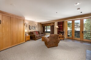 A spacious living room with a large wooden wardrobe, a sofa set, a coffee table, and a chair. There is a large window with a view of the outside. - Ski-In/Out Condo | Lone Peak Views & Community Pool/Hot Tub (Big Sky)