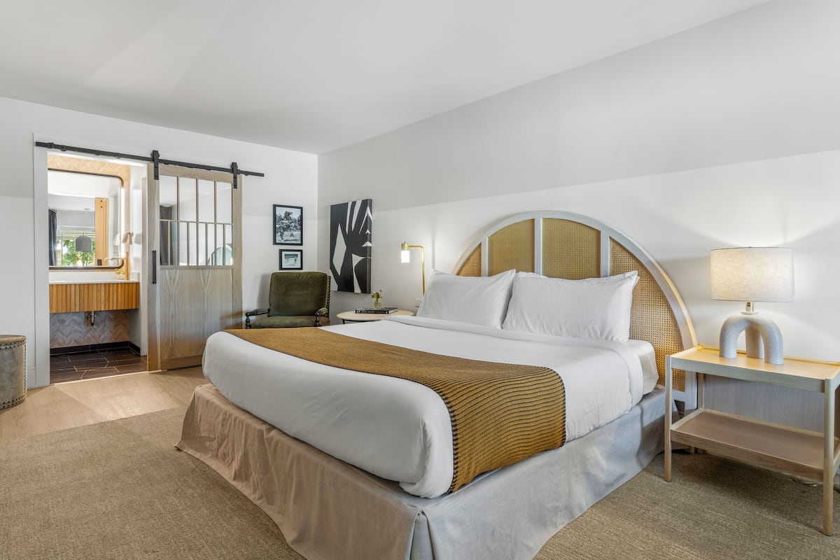 A modern hotel room with a large bed, a bedside table with a lamp, a chair, and a view of the bathroom.