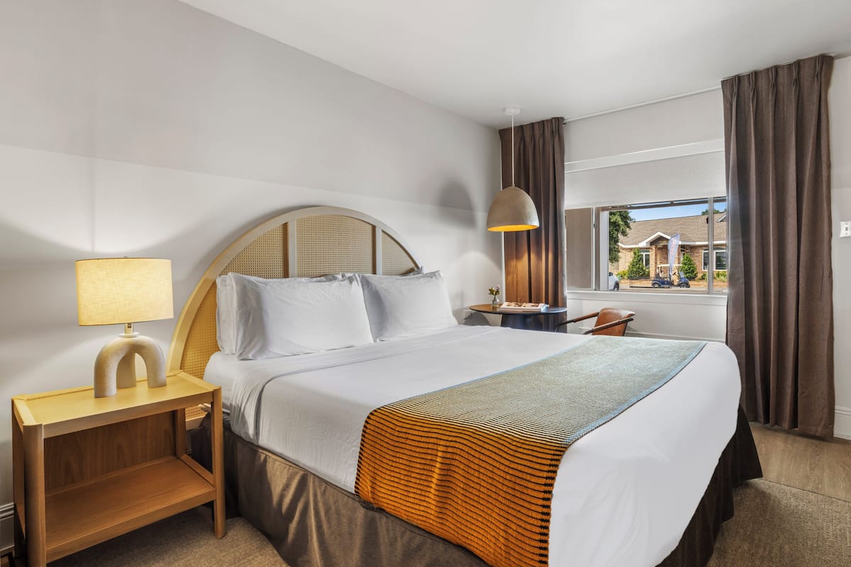 A hotel room with a bed, a nightstand with a lamp, a desk with a chair, and a window with curtains.