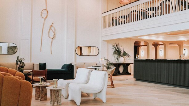 Un vestíbulo espacioso con techo alto, una zona de estar central con sofá y sillones, una recepción y una escalera que conduce a un nivel superior.
