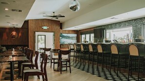 Un restaurante con barra, mesas y sillas de madera, ventilador de techo y un amplio ventanal con vistas a la ciudad.