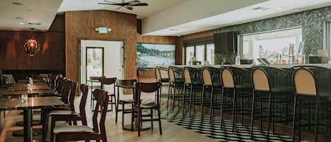 A restaurant with a bar area, wooden tables and chairs, a ceiling fan, and a large window overlooking a cityscape.