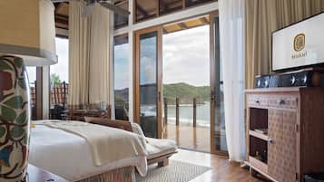 A bedroom with a bed, a TV, a wooden cabinet, and a balcony with a view of the sea.