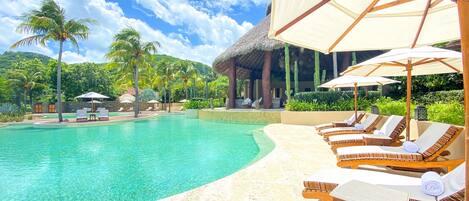 A resort pool with lounge chairs and umbrellas, surrounded by palm trees and a thatched-roof building.