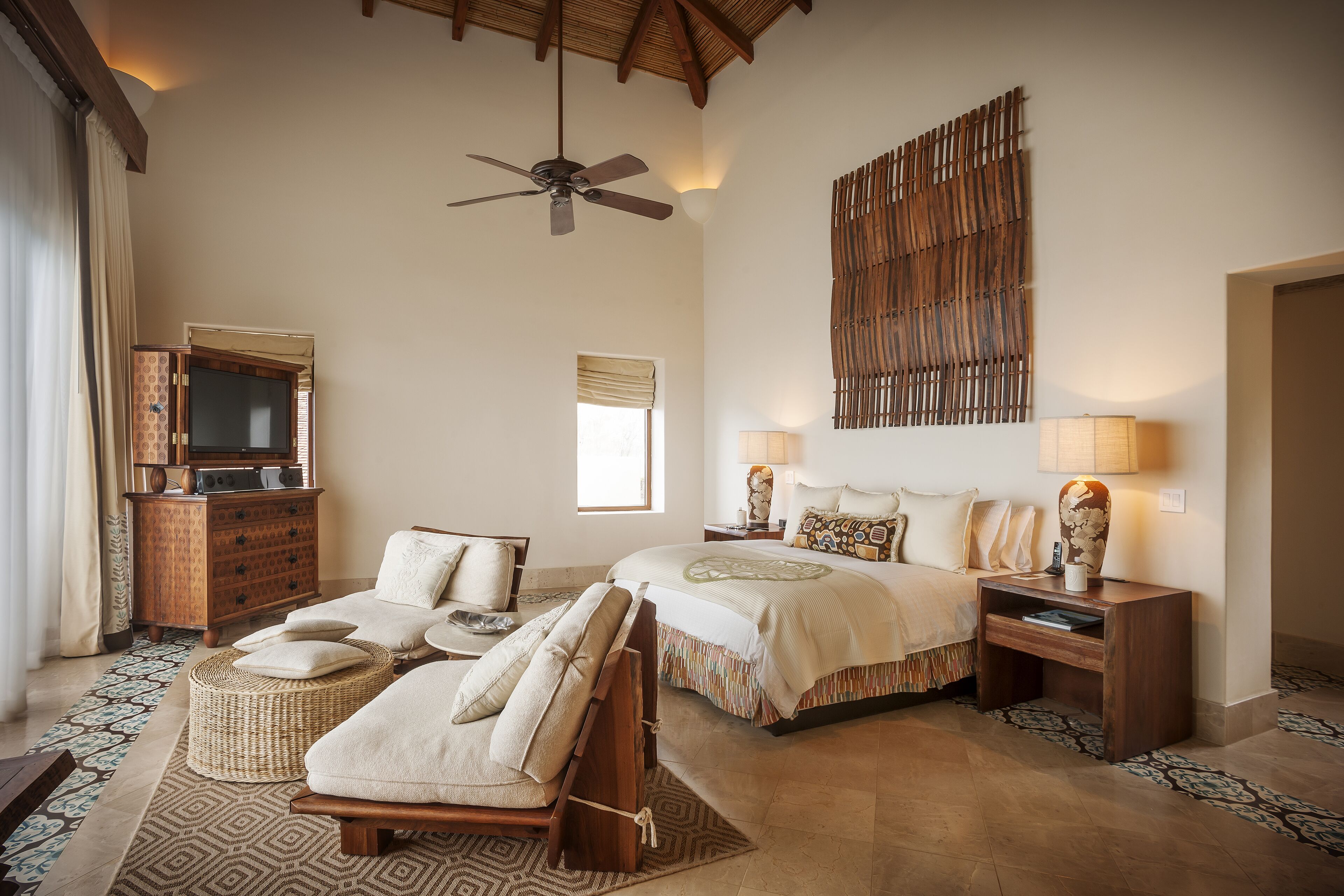 A spacious bedroom with a large bed, two bedside tables, a ceiling fan, a decorative wall panel, and a wooden dresser.