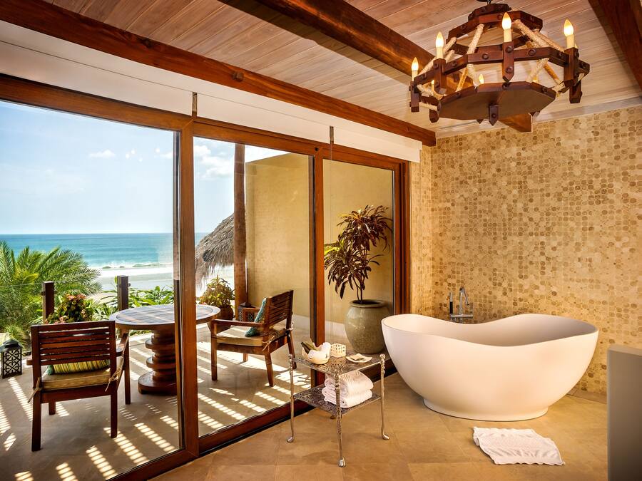 A modern bathroom with a freestanding bathtub, a wooden ceiling, and a large window overlooking the beach.