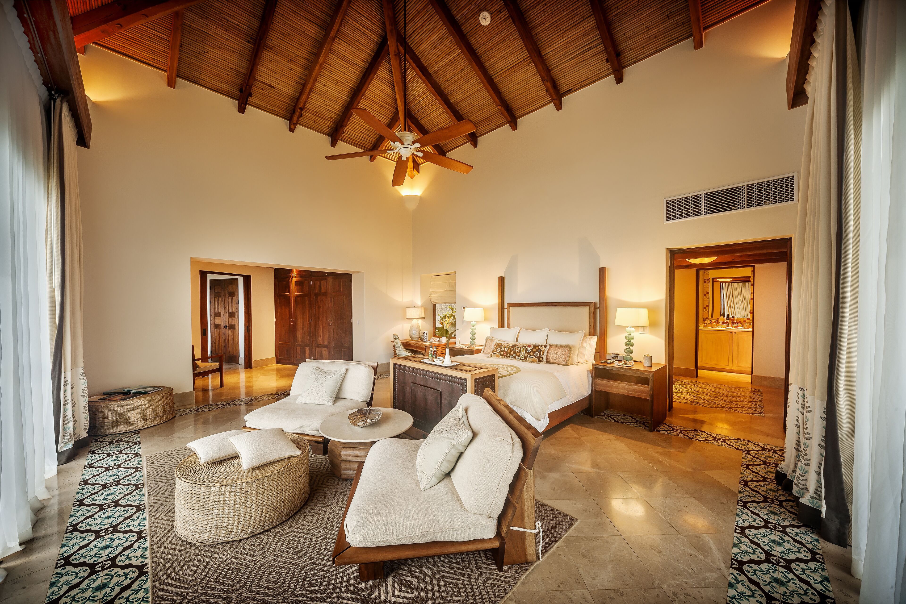 A spacious room with a high ceiling, a ceiling fan, a bed with a nightstand, a seating area with a sofa and chairs, and a patterned rug.