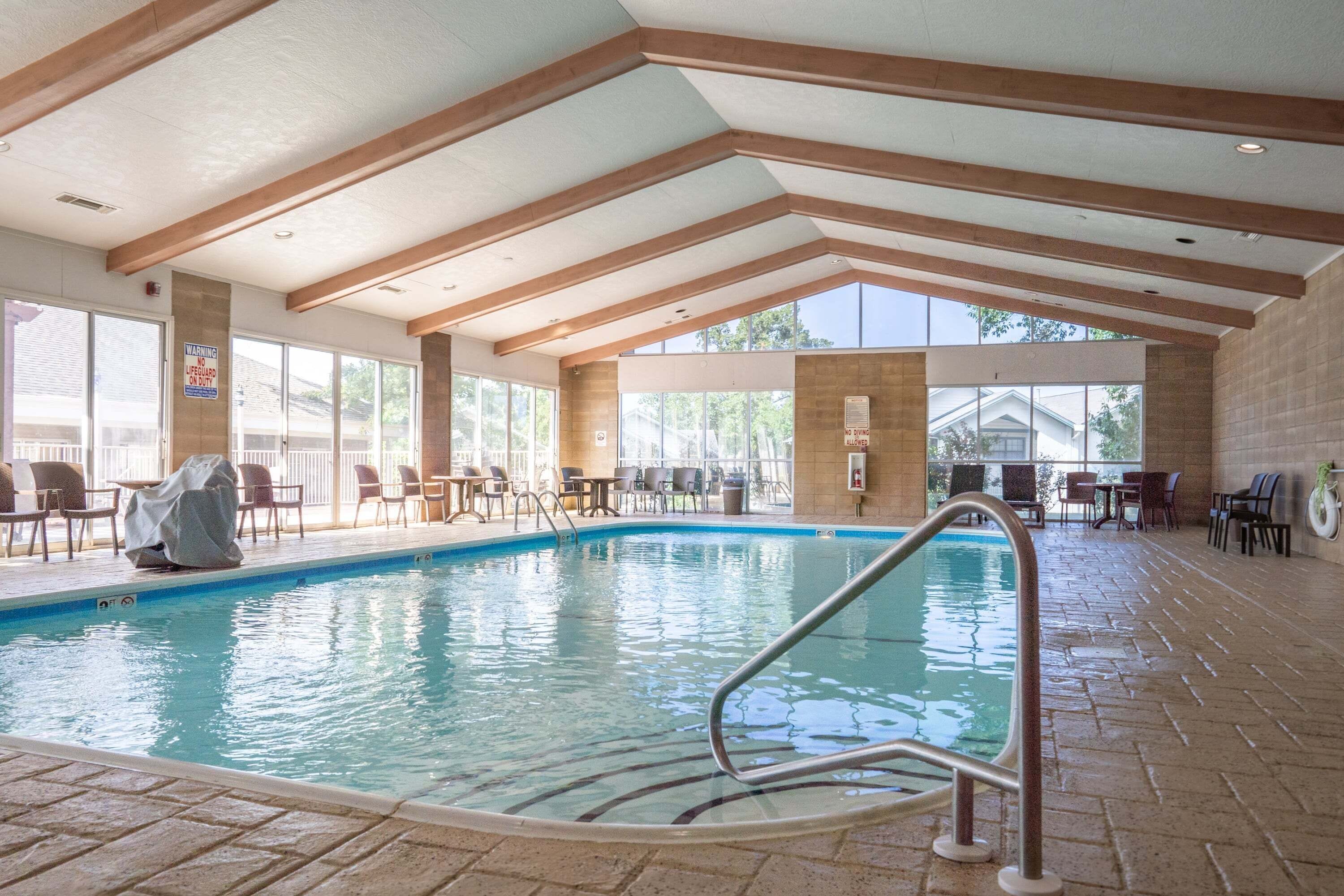 An indoor swimming pool with a clear view of the surrounding area, including a seating area with tables and chairs, and a view of the outside through large windows.